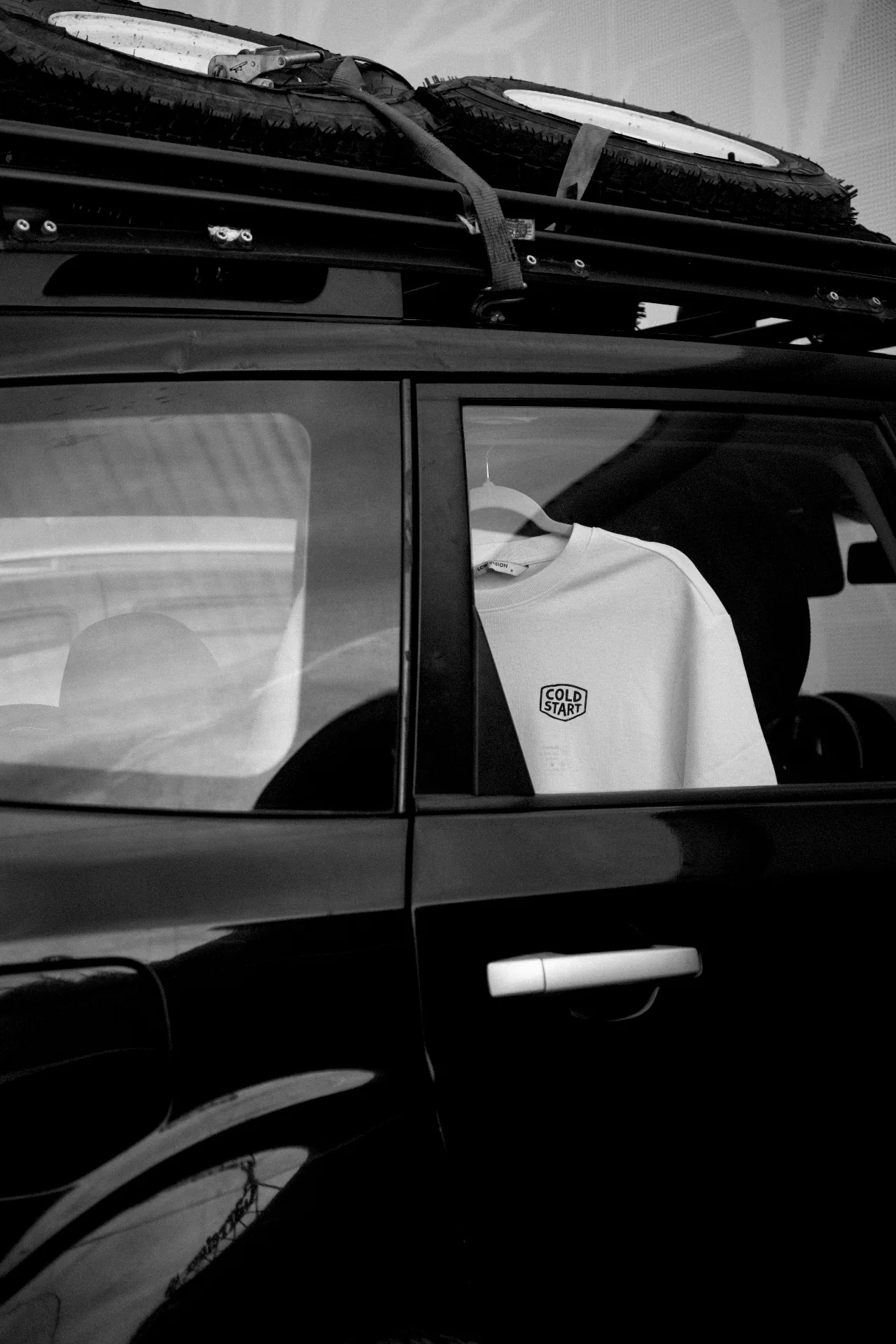 Cold Start tee hanging in a black off-road car with spare tires on the roof rack